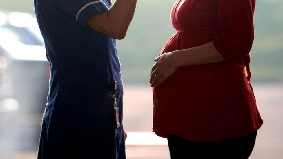 A nurse in a blue uniform talks to a pregnant person in a red top