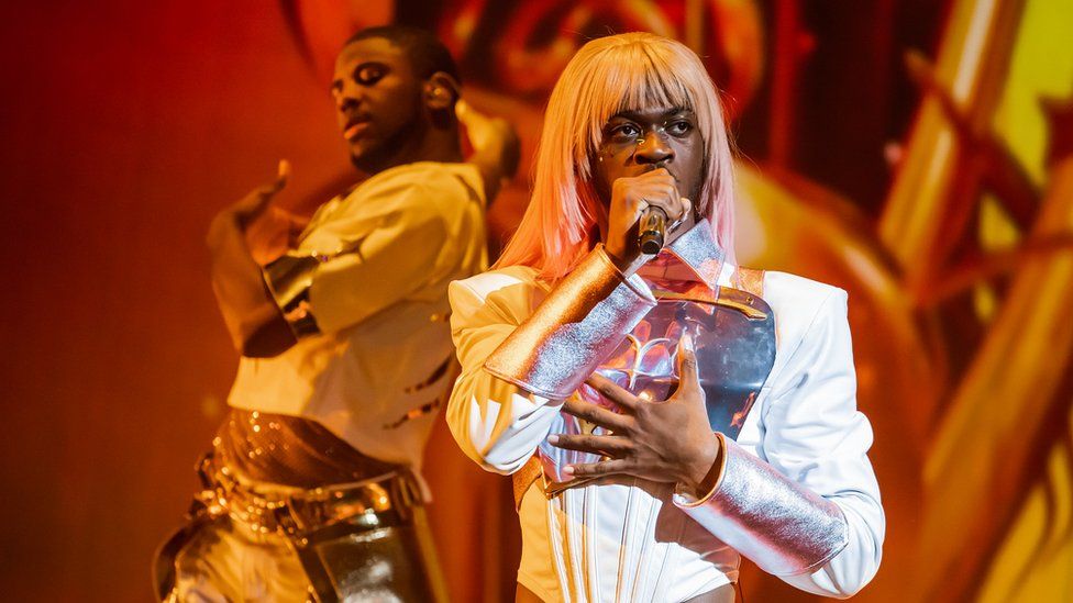 Lil Nas X wears a white jumpsuit and blonde-pink wig. The suit has large silver cuffs from the wrists to the elbows, and a silver chestplate. He's holding a microphone to his mouth and his other hand is pressed, palm flat against his chest. In the background a dancer wearing a similar white outfit but no wig is in the middle of a dance move, one arm outstretched and the other extended behind him. His eyes are closed. The stage is bathed in an orange-gold light.