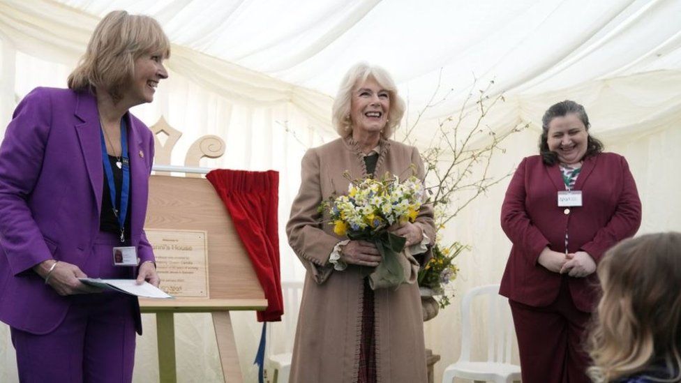 Queen Camilla holds a bouquet of flowers in a marquee in front of a plaque with two staff members next to her