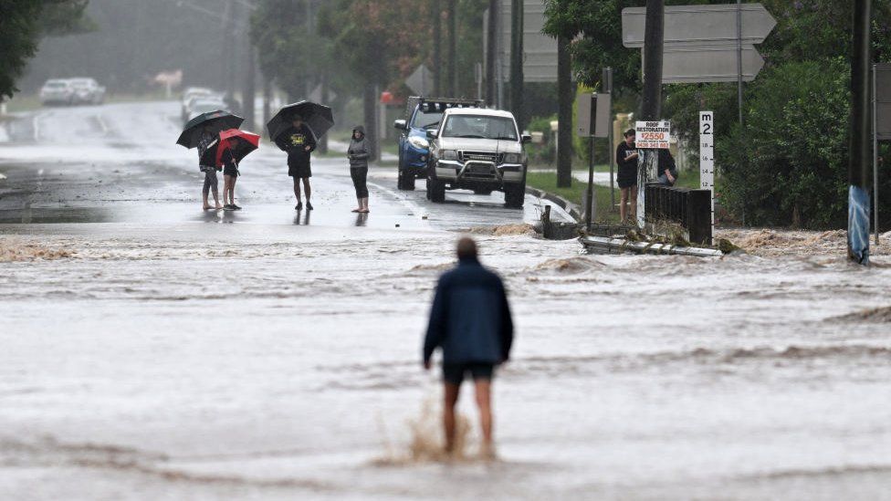 A man wades through floodwater covering a road as people look on