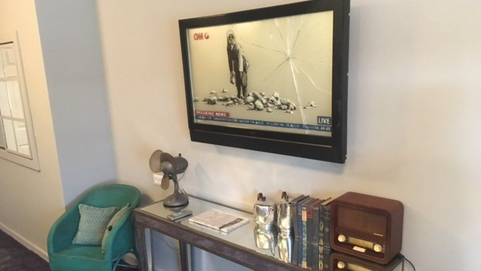 A mock television shows a painting by Banksy - a girl throwing rocks at the camera, apparently on the CNN channel. But the glass of the fake television is itself broken by her assault.