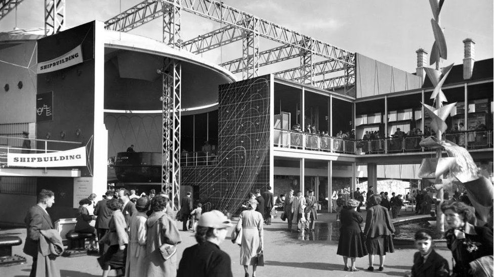 Exterior view of part of the shipbuilding display at the Festival of Britain. On the left is the Power and Production Pavilion and on the right the Natural Resources Pavilion. The 'Skylon' is seen rising from behind the Dome of Discovery in the distance. On the right, is seen part of the Royal Festival Hall and Shot Tower.