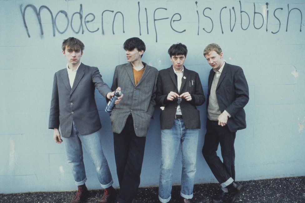 Blur's four members holding spray cans in front of graffiti on a wall reading "Modern life is rubbish"