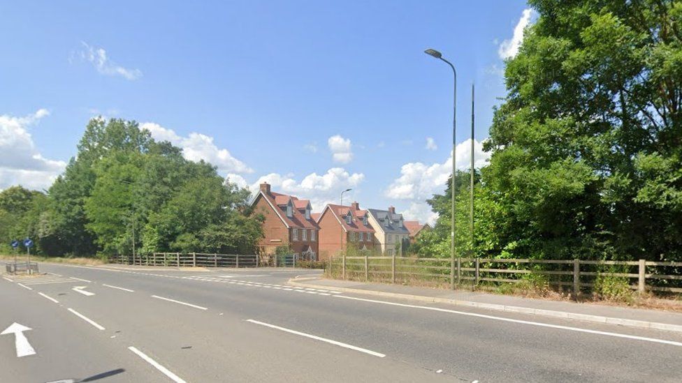 A road and junction on the right, with new houses seen with trees and wooden fencing alongside