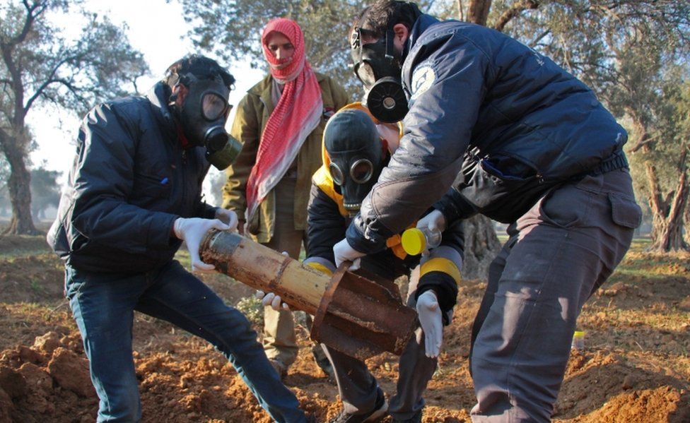 Syria Civil Defence members in gas masks carefully handle remnants of a rocket
