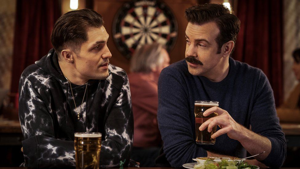 Actor Phil Dunster as his character Jamie Tartt sat in a pub with Jason Sudeikis as his character Ted Lasso. Jamie has a pint of beer in front of him and Ted is holding a pint of beer in his left hand. There's a dartboard in the background behind them