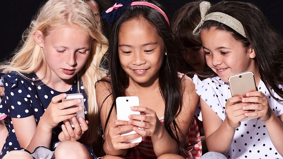 Three primary age girls sitting down looking at their smartphones