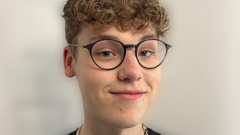 Thomas Fage is a 19-year-old white man with blue eyes and curly dirty blonde hair. He wears round black-rimmed glasses and smiles at the camera. 