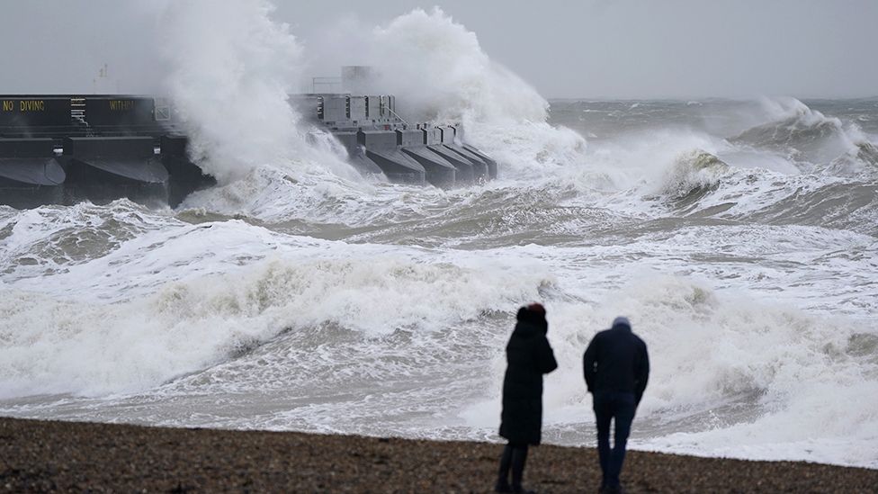 Two people silhouetted on a beach looking out to sea as strong winds and waves hit a pier on 31 December 2023
