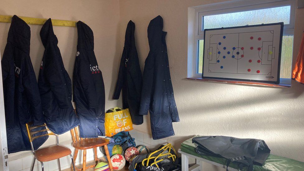 Coats hanging on hooks in the corner of a room with balls and other football equipment on the floor