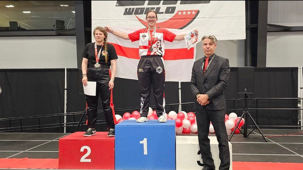 Teenage girl stands on centre podium above the number 1. Another competitor stands to her right above 2.