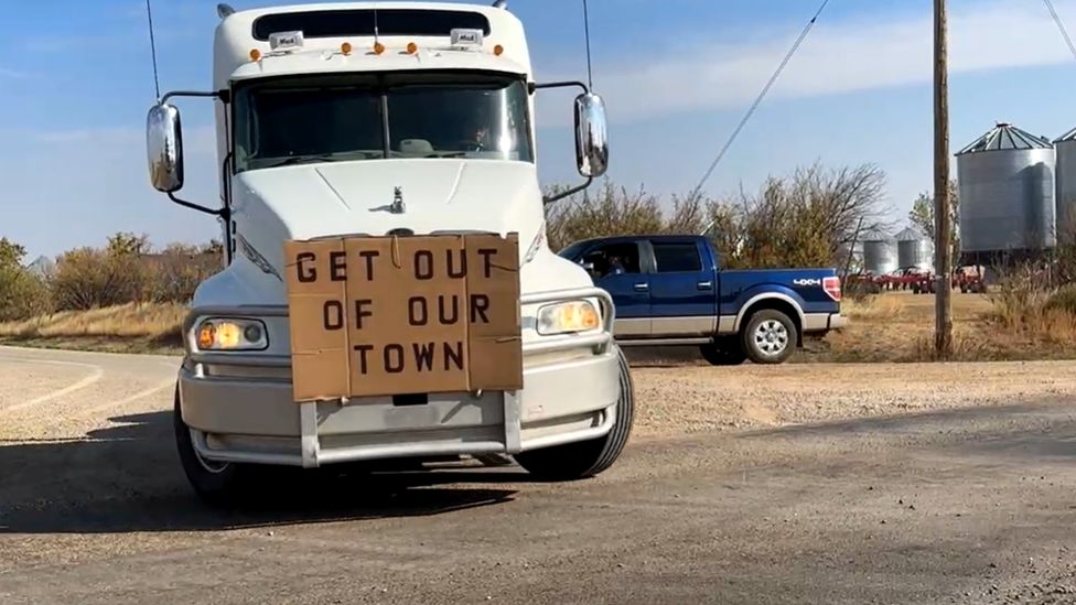 White semi-truck with cardboard sign reading Get out of our townin front of blue truck on road.