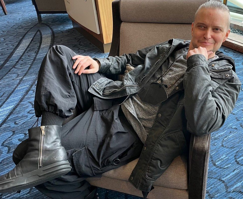 Lucien Greaves smiles while lounging in a chair at the Marriott Copley hotel during SatanCon. He's wearing a hoodie and a black Barbour jacket.