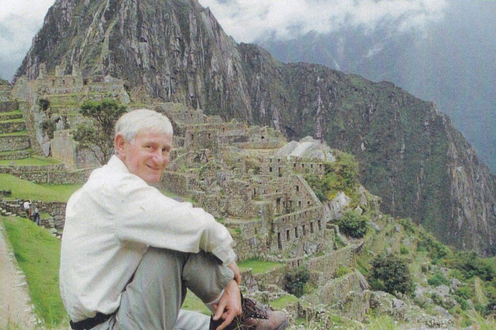 Bill sits on a hill overlooking old ruined buildings with a mountain beyond
