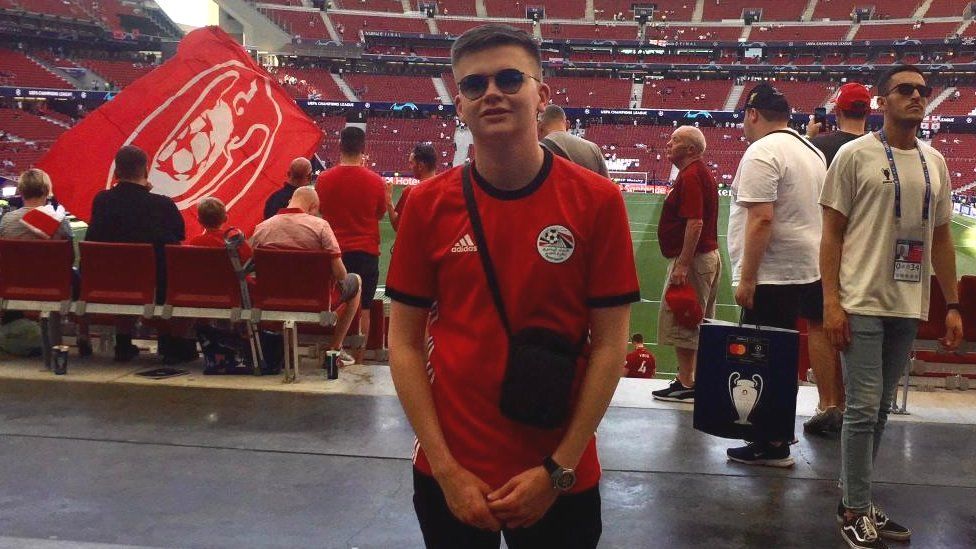 Josh wearing sunglasses, a bag and a red Egypt shirt at Wembley Stadium with other fans in the background.