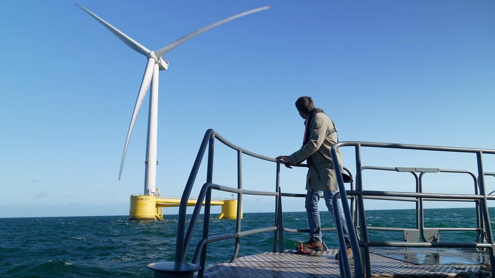 Climate editor Justin Rowlatt holding railing on back of boat looking at floating wind turbine