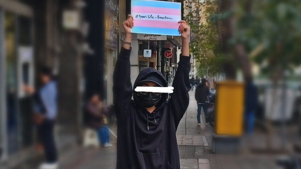 A young Iranian LGBTQ person holds up a trans flag reading "queer, life, freedom"