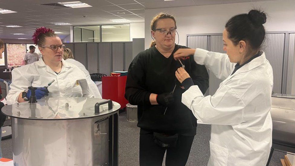 Two women in lab coats and a third woman dressed in black work on a washing machine