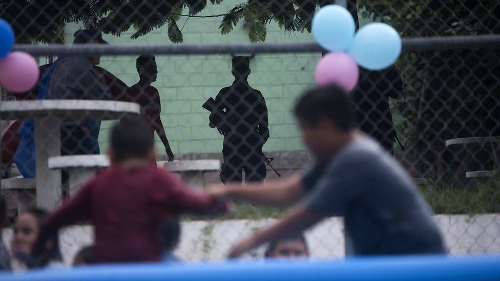 Children playing on a concrete court, while armed youths watch on