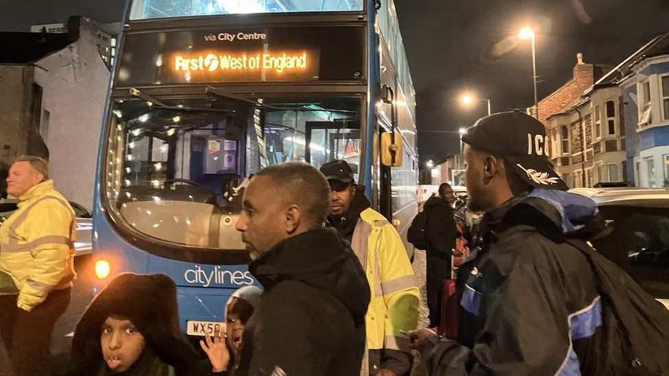 A bus parked on a city street at night with people waiting in front of it