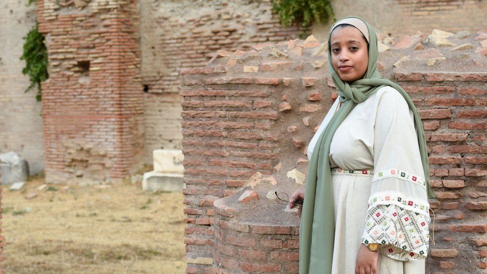 Kenyan writer and poet Khadija Abdalla standing on a historical site, there are ruins of brick buildings behind her. She is wearing a green headscarf and a white dress.