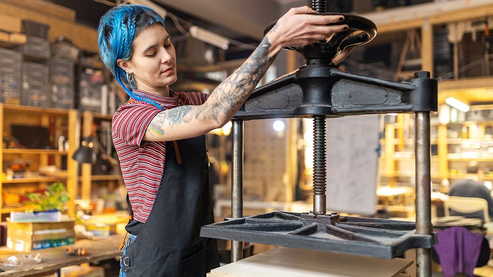 Person with blue hair and tattoos works at a metal press in a workshop