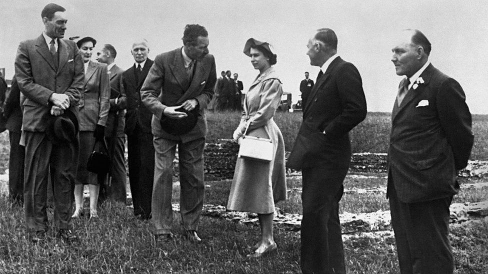 The Queen with men in suits on a small hill