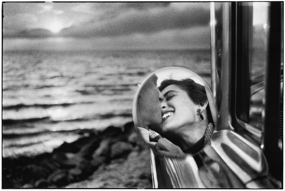 A couple is seen kissing through the rear-view mirror of their car, which is parked facing the sea, California, 1955