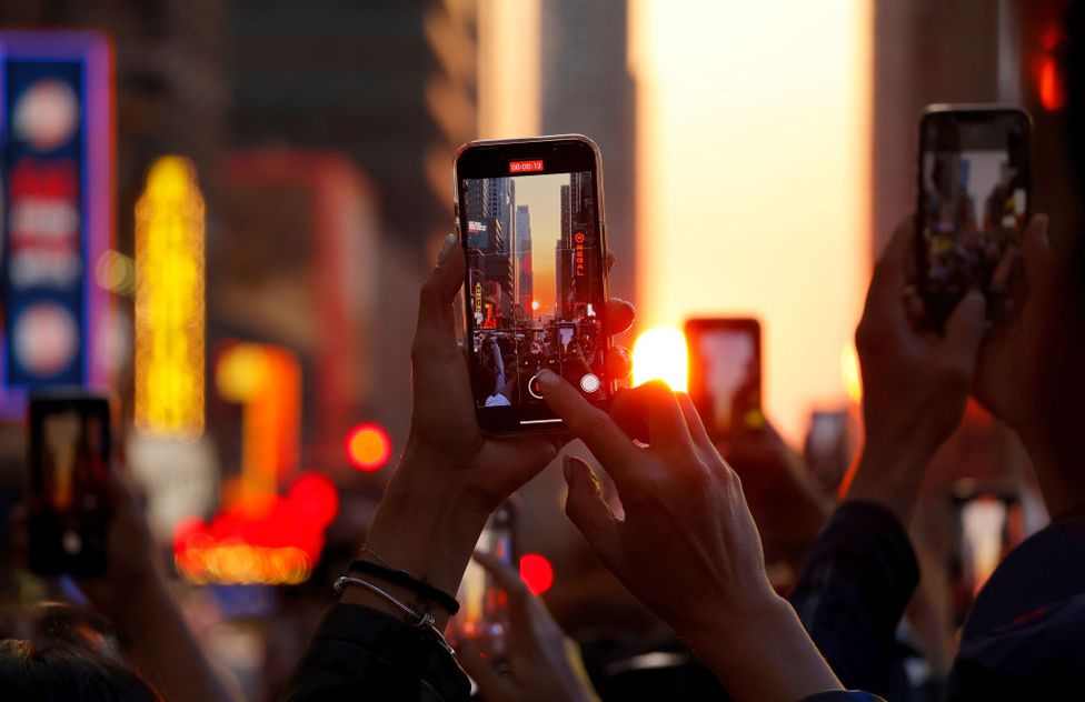 People stand on 42nd Street in Times Square as they photograph the second evening of the Manhattanhenge sunset on May 30, 2023, in New York City.