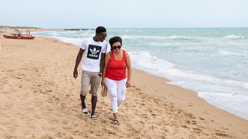 Muhammed Sanneh and Giusella Ferraro walking on a beach in Sicily