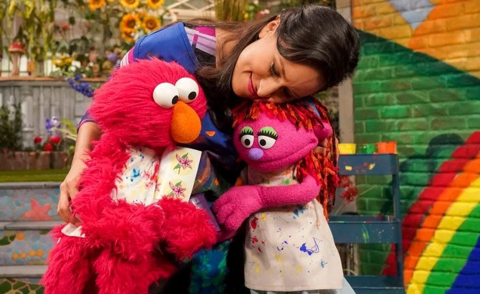 An actress in Sesame Street hugs the muppets Elmo and Lily