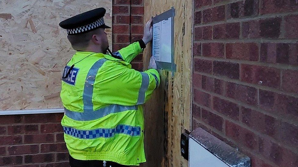 Police officer in hi-viz fixes a notie to a boarded up house
