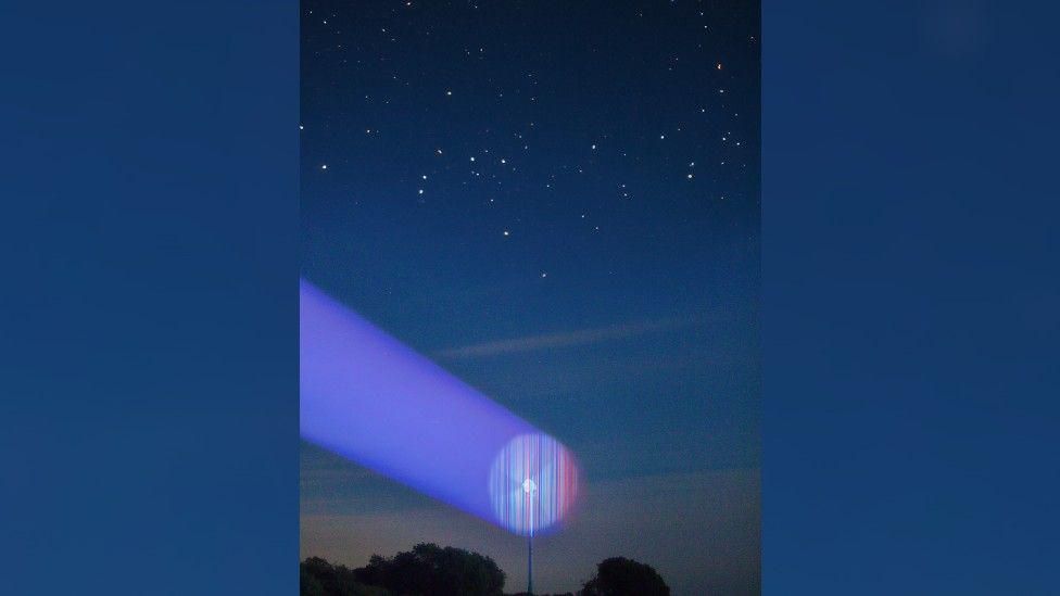 A wind turbine with a circular version of the blue-to-red climate stripes projected into the arc of its blades, underneath a starry sky