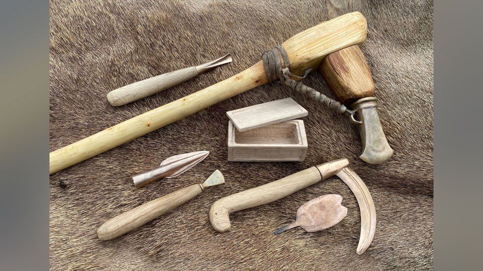 Four wooden handled replica Bronze Age tools, plus a replica Bronze Age box, arrow head and blade against a fur background