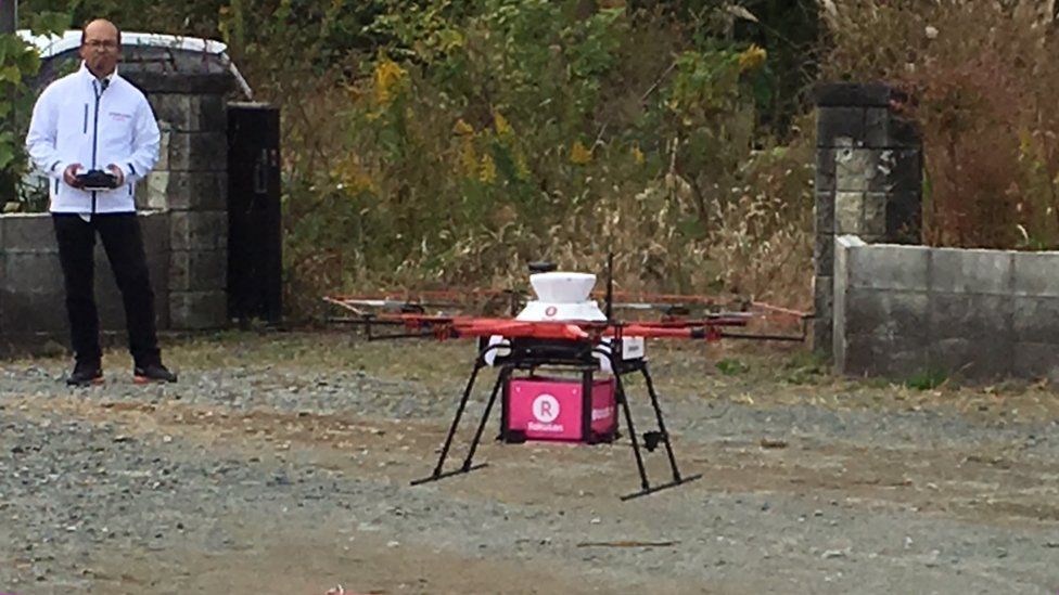 A man stands behind a drone sitting on the ground, which can deliver food to people in Japan