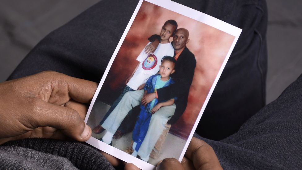 Moti Dereje holding a family photo showing himself as a boy (L), his father (C) and his younger brother (R)