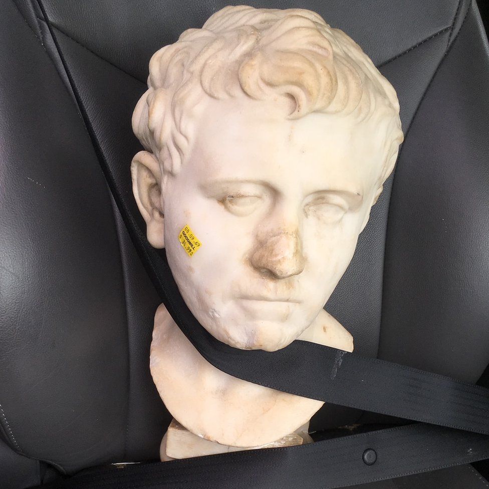 A Roman bust is buckled in the passenger seat of a car.