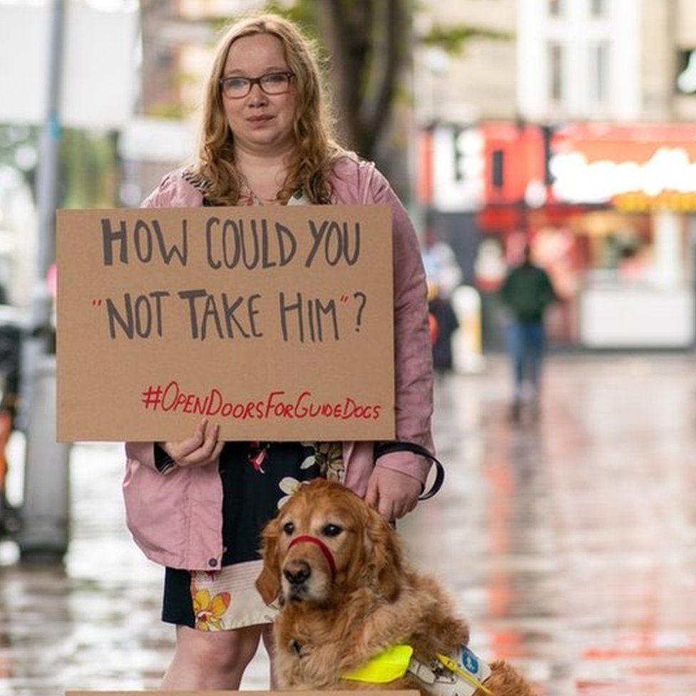 Angharad Paget-Jones holding a placard saying 'how could you refuse him?' next to her guide dog Tudor