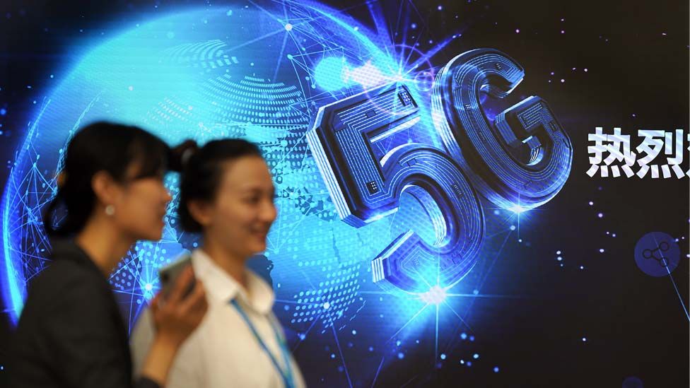 Two women walk in front of an LED wall displaying the words "5G, Warm Welcome" in at China Mobile Office in Beijing, China
