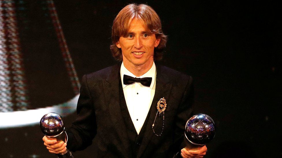 Luke Modric is seen in a tuxedo, holding two awards - one in each hand - in the middle of an awards ceremony