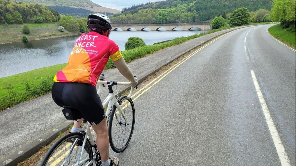 Judith cycling, wearing a Breast Cancer Now t-shirt at Ladybower Reservoir in Derbyshire.