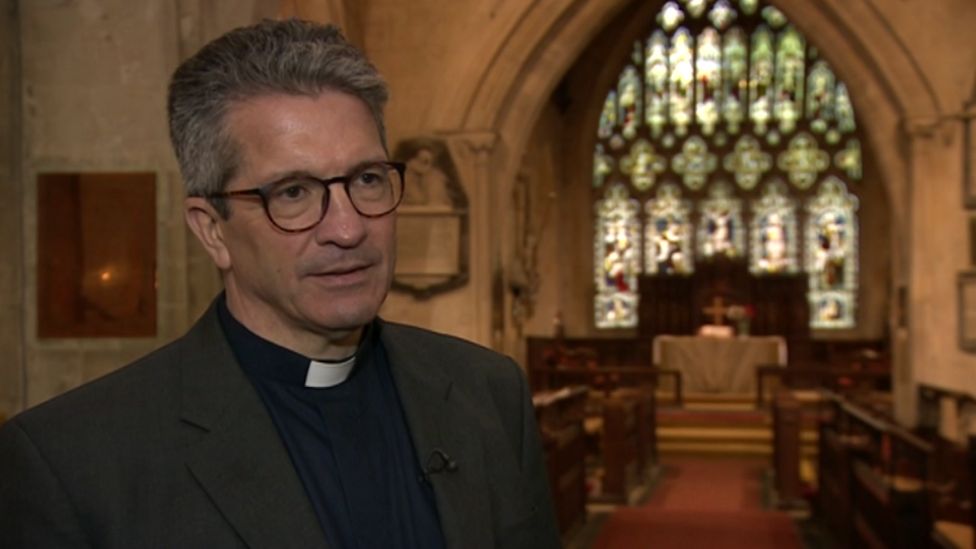 A vicar with grey hair in a vicars collar and black outfit and black glasses