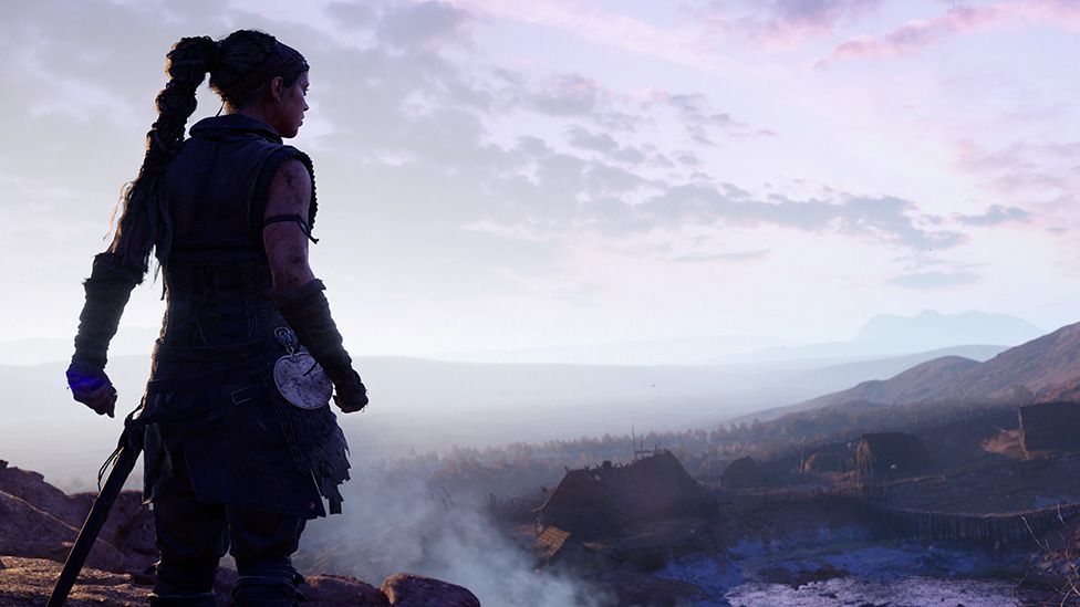A female figure with a large sword sheathed at her side stands silhouetted against the horizon as she looks out over a sprawling, hilly landscape with wooden huts dotted around it. Mist rises from the ground and the purple hued sky suggests a dawn scene.