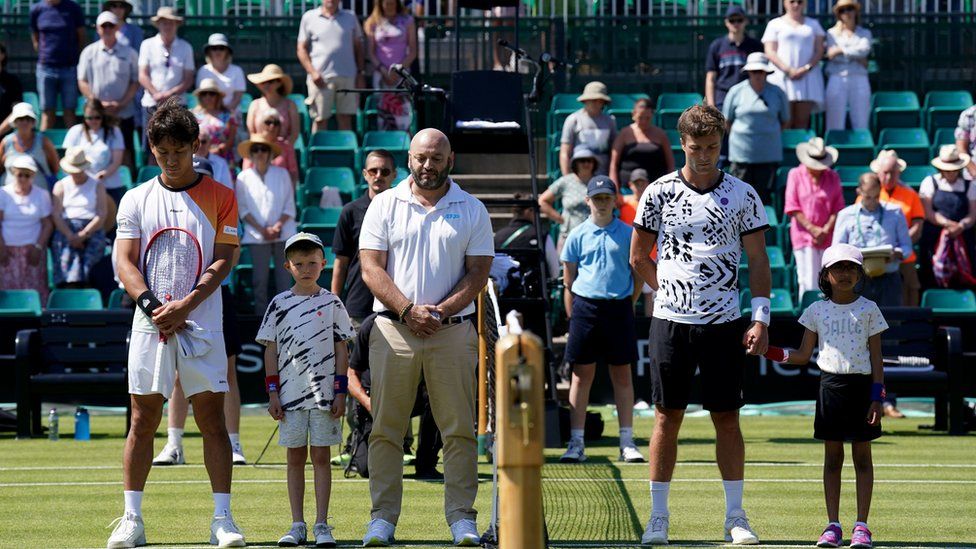 Sho Shimabukuro (left) and Liam Broady stand with the umpire, mascots and fans for a minute's silence in memory of the victims of yesterday's attacks in Nottingham on day three of the Rothesay Open 2023 at the Nottingham Tennis Centre.