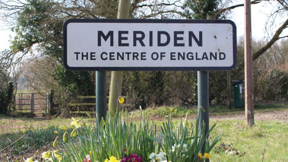 Sign with black writing on a white background with daffodils underneath. It reads "Meriden The Centre of England" 