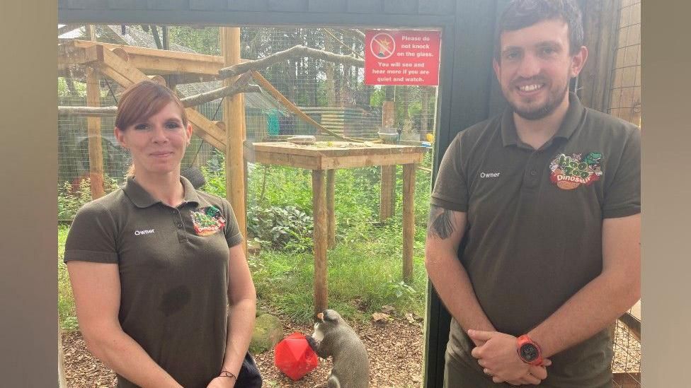 A man and woman stand in front of a glass window that leads to a monkey enclosure. The woman has auburn hair that is tied up and she is wearing a brown T-shirt. She is standing on the left. The man, standing on the right, has brown hair, and is wearing a similar brown T-shirt. In the enclosure behind them is green grass and brown wood chippings on the floor. There is brown wooden climbing platforms for monkeys. At the bottom of the frame, on the floor of the enclosure, is a brown monkey holding a red ball.