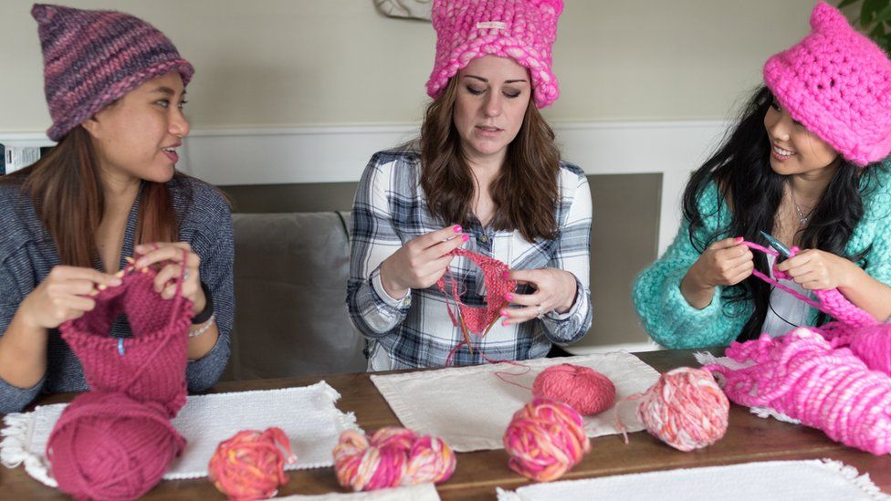 Three woman, wearing pussyhats, are seen talking to each other as they knit more at a table piled with balls of pink yarn