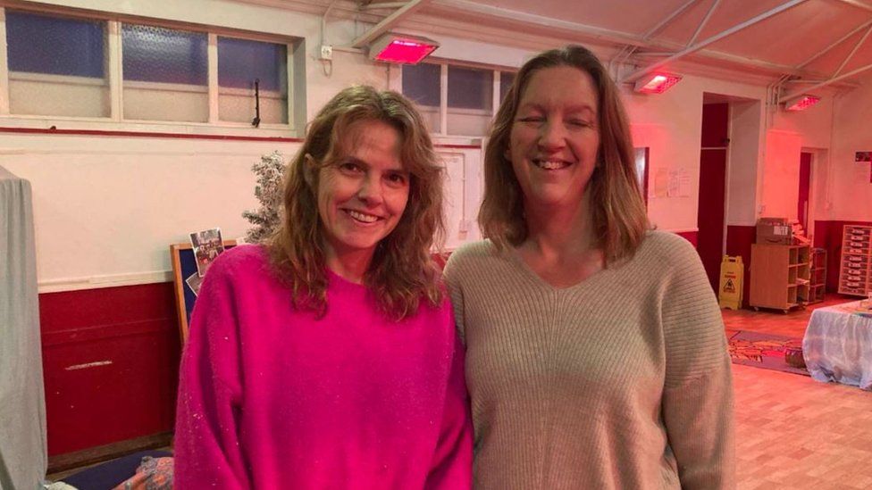 Two white women with long dark blonde hair wearing pink and grey jumpers smile at the camera