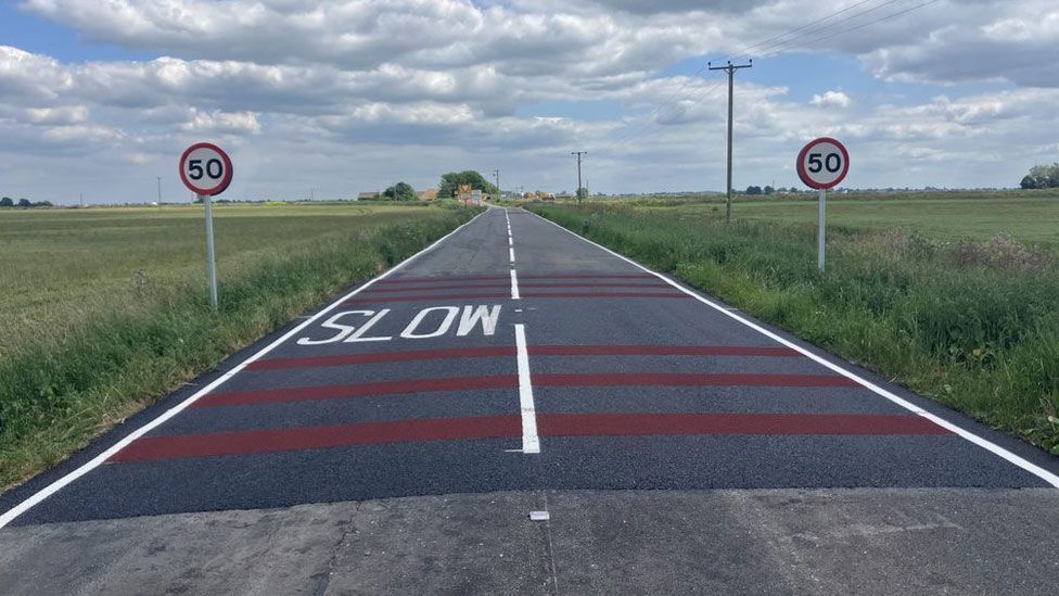 A road with 50mph signs on either side and slow in white letters written on black tarmac with red countdown markers across