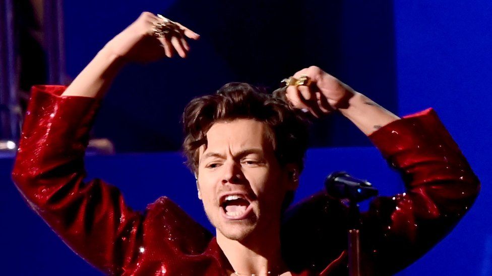 Harry Styles performs astatine  the Brit Awards. The signifier    down  him is lit up   successful  purple, and he's wearing a glittery burgundy jacket. He has some  hands raised supra  his head, astir   and looks to beryllium  successful  the mediate  of singing a note, and wears golden  rings connected  his fingers.